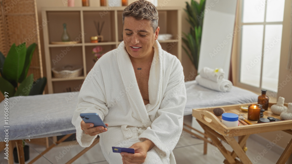 Young man in a spa wearing a white robe, holding a phone and card, surrounded by wellness items in an indoor setting, conveying relaxation and modern lifestyle.