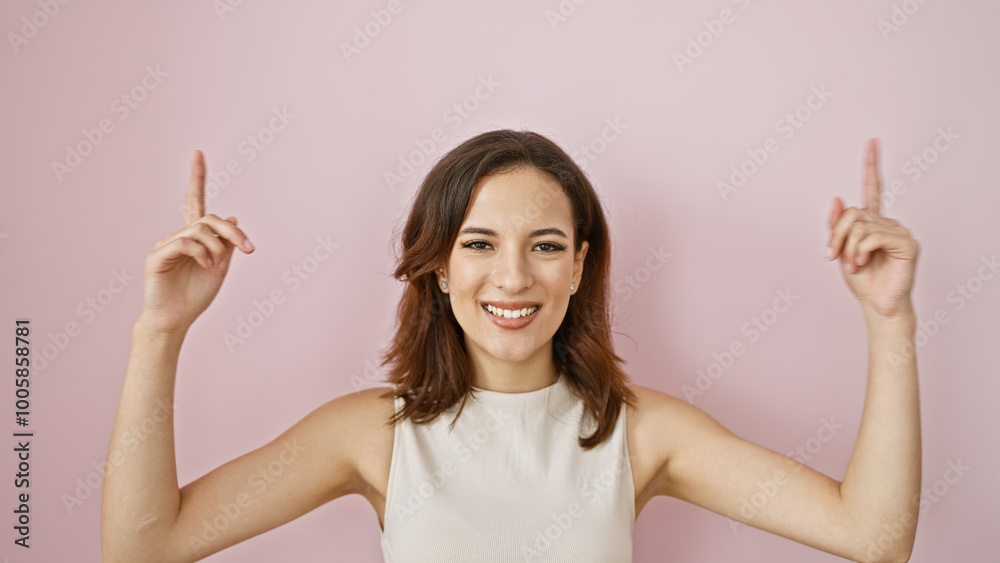 A smiling young hispanic woman with a beautiful hairstyle points upwards, isolated against a pink wall background. A smiling young hispanic woman with a beautiful hairstyle points upwards, isolated against a pink wall background.