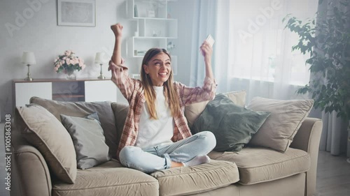 Young woman sitting on a couch in a cozy living room celebrates a victory while playing an online game on her smartphone, radiating happiness and excitement