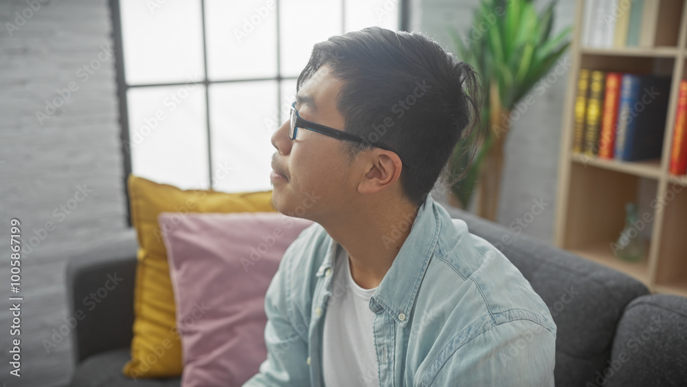 Pensive young asian man with glasses looking away while sitting on a sofa in a cozy living room. Pensive young asian man with glasses looking away while sitting on a sofa in a cozy living room.