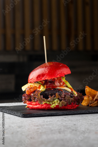 A gourmet burger with a striking red bun features fresh toppings like lettuce, avocado, and bacon. The burger is served steaming hot on a black slate with a wooden skewer.
