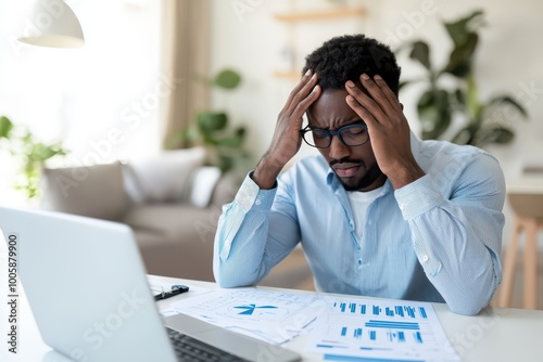 A stressed man with glasses is sitting at a desk, struggling with reports and data on his laptop, reflecting workplace pressure and frustration.