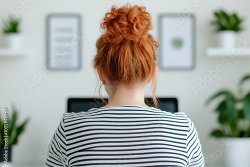 A woman with her hair in a bun, working on a computer in a cozy home office setup with potted plants.