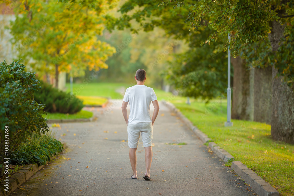 Φωτογραφία Young adult man in white t-shirt and shorts walking on asphalt road at city park in beautiful warm summer day