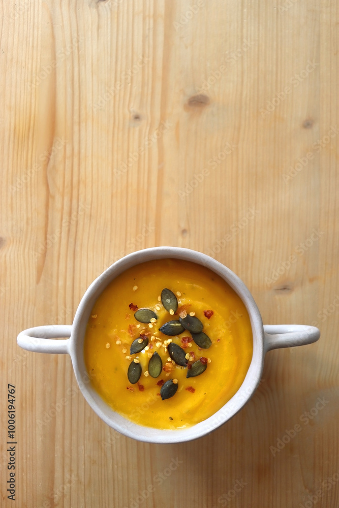 Bowl of creamy pumpkin and potato soup, with pumpkin seeds and chilli flakes toppings. Top view, wooden background. Bowl of creamy pumpkin and potato soup, with pumpkin seeds and chilli flakes toppings. Top view, wooden background.