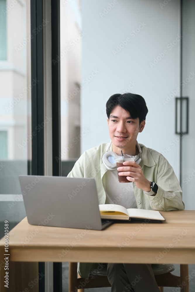A Young Asian man is savoring a cup of coffee at a table and looking out of the window, blending relaxation with inspiring for a perfect start to the day A Young Asian man is savoring a cup of coffee at a table and looking out of the window, blending relaxation with inspiring for a perfect start to the day