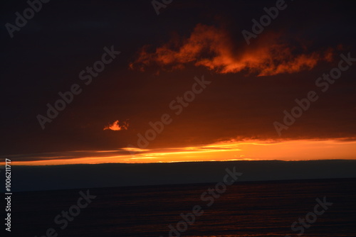 A stunning sunset over the ocean with dramatic dark clouds and a vivid orange and red horizon, casting warm reflections on the calm water.