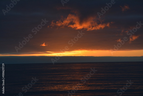 A stunning sunset over the ocean with dramatic dark clouds and a vivid orange and red horizon, casting warm reflections on the calm water