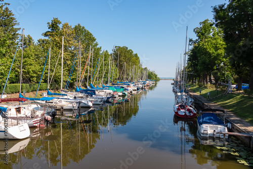 Picturesque marina with line of sailboats docked along narrow channel in Balatonmariafurdo at Balaton lake, Hungary. Colorful sails and masts. Vibrant nautical atmosphere. Reflection on calm water photo