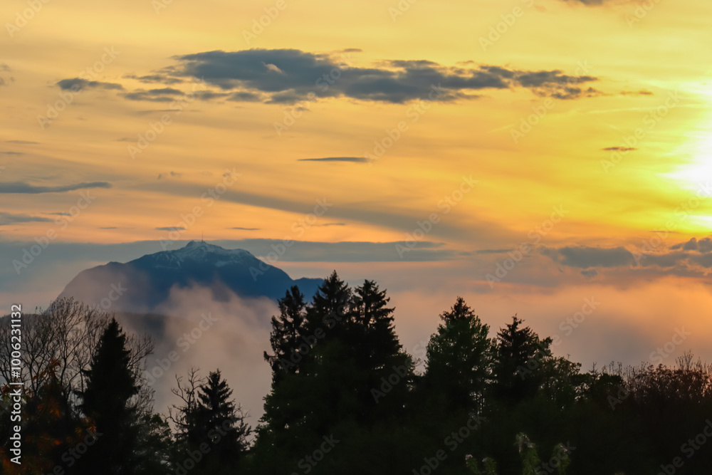Fiery sunset over mountain summit Dobratsch in Rosental, Carinthia, Austria, Europe. Silhouette of forest and rolling hills in Austrian Alps. Vibrant orange and yellow hues. Tranquil serene atmosphere Fiery sunset over mountain summit Dobratsch in Rosental, Carinthia, Austria, Europe. Silhouette of forest and rolling hills in Austrian Alps. Vibrant orange and yellow hues. Tranquil serene atmosphere