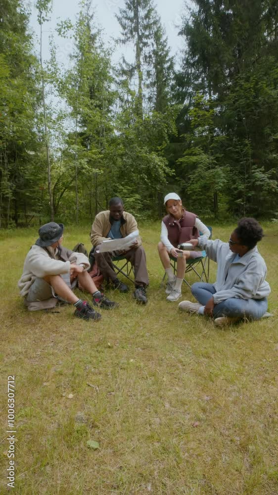 Vertical shot of four multiethnic male and female hikers resting on meadow and exploring paper map while on hiking trip in forest setting Vertical shot of four multiethnic male and female hikers resting on meadow and exploring paper map while on hiking trip in forest setting