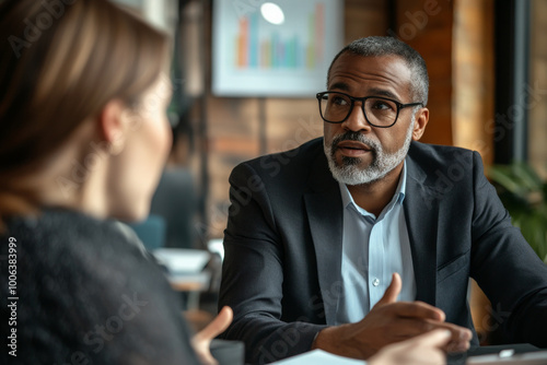 A senior Hispanic executive listens attentively as a middle-aged Black team leader discusses risk transfer strategies. The meeting room is filled with charts and visual aids to enhance understanding. photo