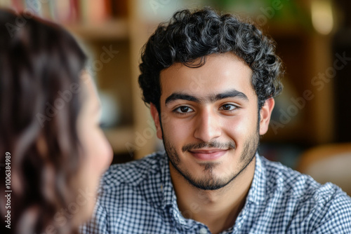 A close up of a young adult Arabian man interviewing a Brazilian woman, both displaying expressions of curiosity and engagement. The background provides a hint of a cozy office setting, adding context