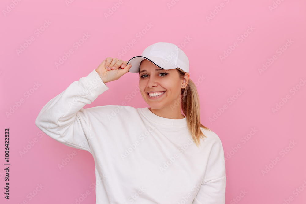 A young happy smiling caucasian beautiful blonde woman in a white sweatshirt and cap standing holding on to the visor of her cap isolated on a light pink color background A young happy smiling caucasian beautiful blonde woman in a white sweatshirt and cap standing holding on to the visor of her cap isolated on a light pink color background
