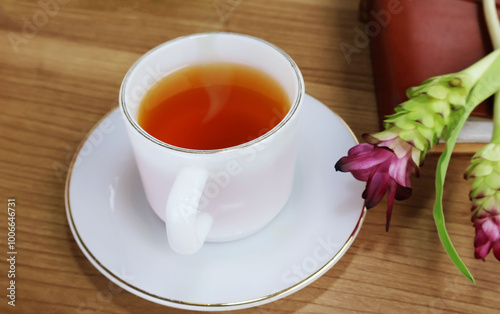 Hot tea in a pearl cup with a saucer on the side. A book with a long bouquet of red and green flowers on a wooden table.