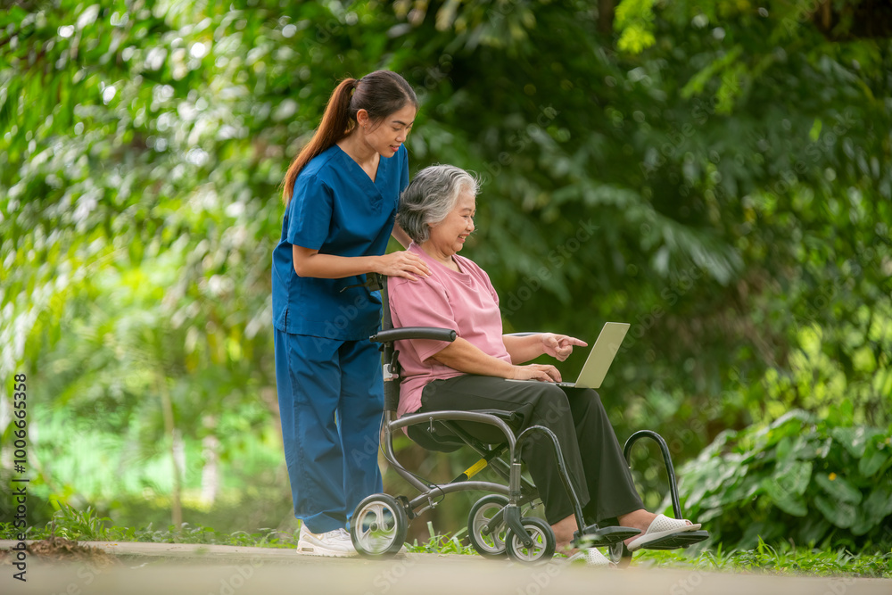 Elder woman is sitting a wheelchair holding a tablet, while a young healthcare professional standing smiling look tablet likely explaining something .They are outdoor park healthy treatment concept. Elder woman is sitting a wheelchair holding a tablet, while a young healthcare professional standing smiling look tablet likely explaining something .They are outdoor park healthy treatment concept.