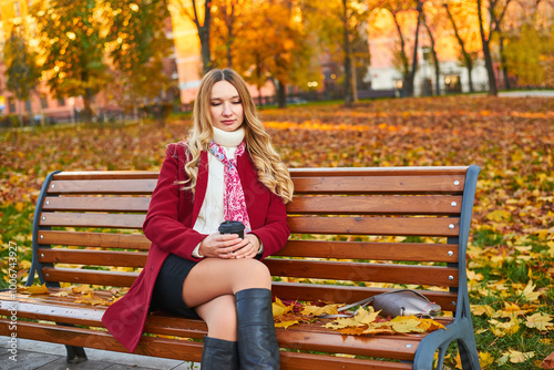 A beautiful woman in a red coat sits on a bench in an autumn park. She is wearing black boots and a scarf.