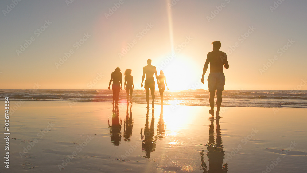 Fototapeta premium Group Of Friends In Swimwear Walk Towards Waves At Sunset On Summer Beach Vacation With Lens Flare