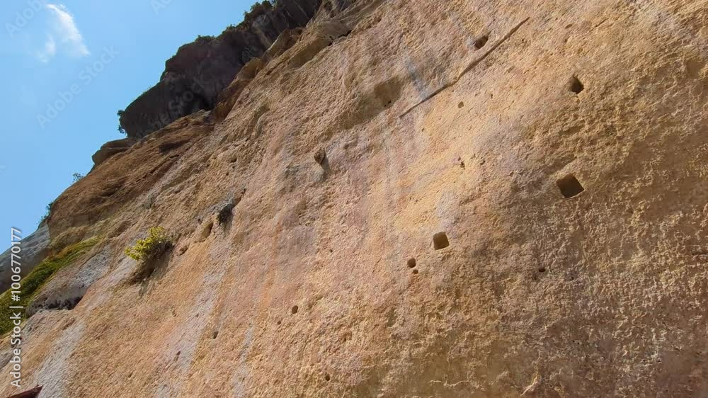 Looking Up View of Imposing Sheer Cliff Historic Rock with Small Man Made Caves and Blue Sky Background 4K