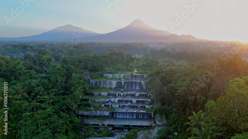 Aerial view of waterfall in the middle of forest in the morning. Mountain and Sun rising on the background. Watu Purbo waterfall, Mount Merbabu and Merapi Volcano. photo