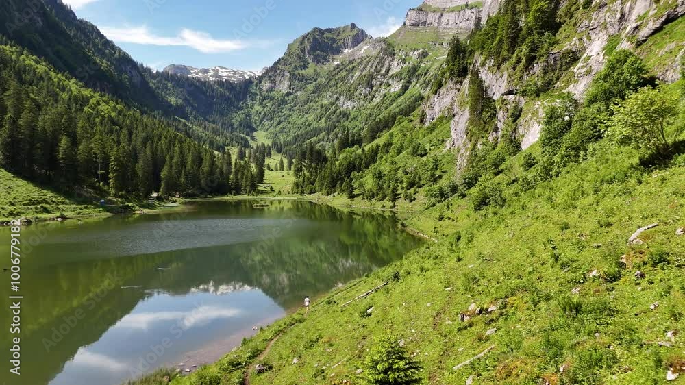 A woman hikes beside a tranquil alpine lake surrounded by towering mountains and lush greenery A woman hikes beside a tranquil alpine lake surrounded by towering mountains and lush greenery