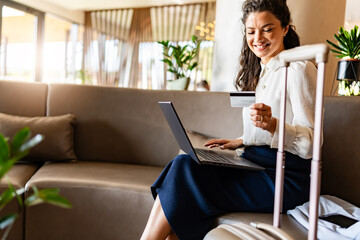 A woman sits on a couch in a modern lobby with her laptop and luggage. She smiles while holding a credit card, ready to make an online purchase or booking.