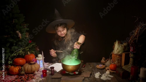 Halloween, smiling little cute girl dressed as witch in witch's hat brews a glowing magic potion in cauldron on coals. Magic wand, сandles, pumpkins, ingridients, fog smoke, magic, mystical atmosphere