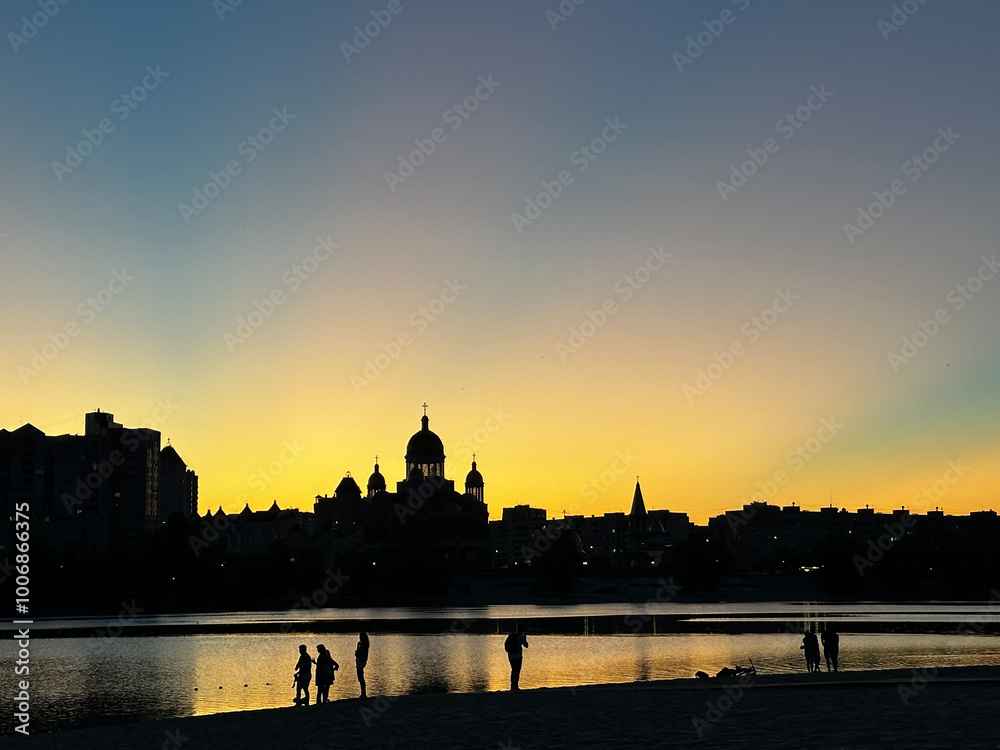 Obraz premium peaceful sunset by a river, with silhouettes of buildings, including a church with domes, against a golden sky. People stand by the water’s edge, enjoying the serene evening scene.