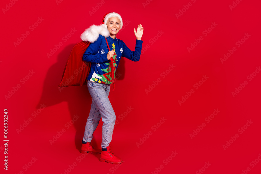 Full body portrait of pretty aged lady gifts bag walk empty space wave hi new year xmas isolated on vibrant red color background Full body portrait of pretty aged lady gifts bag walk empty space wave hi new year xmas isolated on vibrant red color background