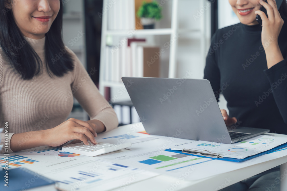 Focused Collaboration: Two professional women work intently at a desk, one using a calculator while the other speaks on a phone, demonstrating a dynamic and productive business partnership. Focused Collaboration: Two professional women work intently at a desk, one using a calculator while the other speaks on a phone, demonstrating a dynamic and productive business partnership.