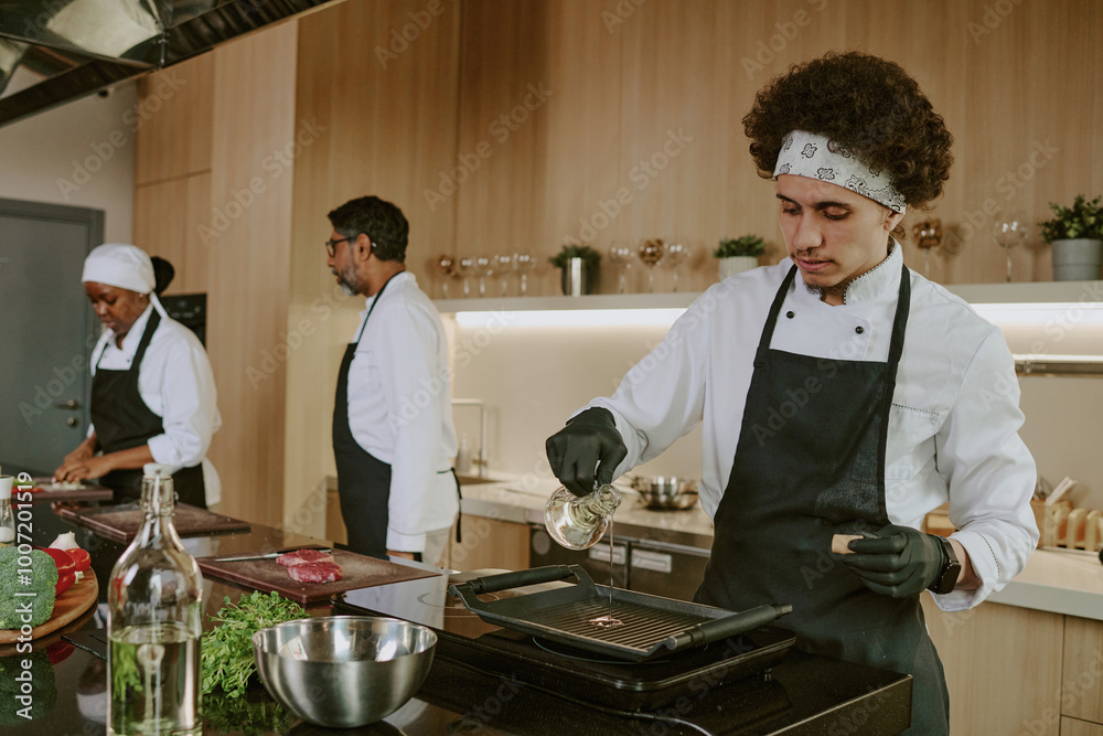 Curly haired sous chef adding olive oil to fry steak, his colleagues cooking in background Curly haired sous chef adding olive oil to fry steak, his colleagues cooking in background