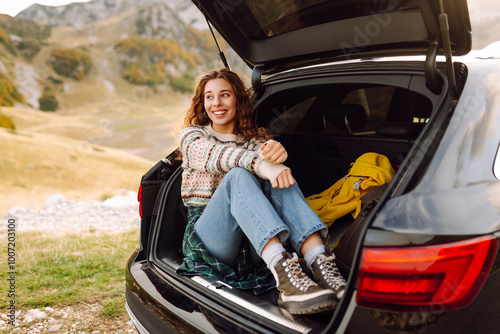 Young woman sits in the trunk of her car, parked on the side of a winding road that cuts through the rocky terrain. Road trips. Autumn landscape