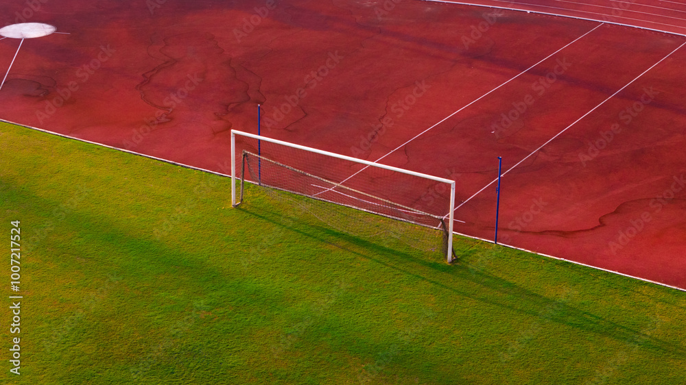 Fototapeta premium A dynamic aerial view of a soccer goal post on a lush green field adjacent to a vibrant red running track, capturing the essence of outdoor sports and athletics