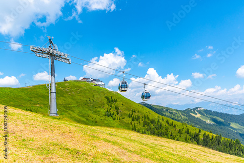 Visitors ride the Schmittenhohe cableway, enjoying stunning views of the Austrian Alps. The vibrant green landscape stretches below under a bright blue sky dotted with clouds. photo