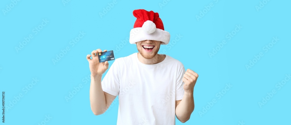 Happy young man in Santa hat and with credit card on light blue background Happy young man in Santa hat and with credit card on light blue background