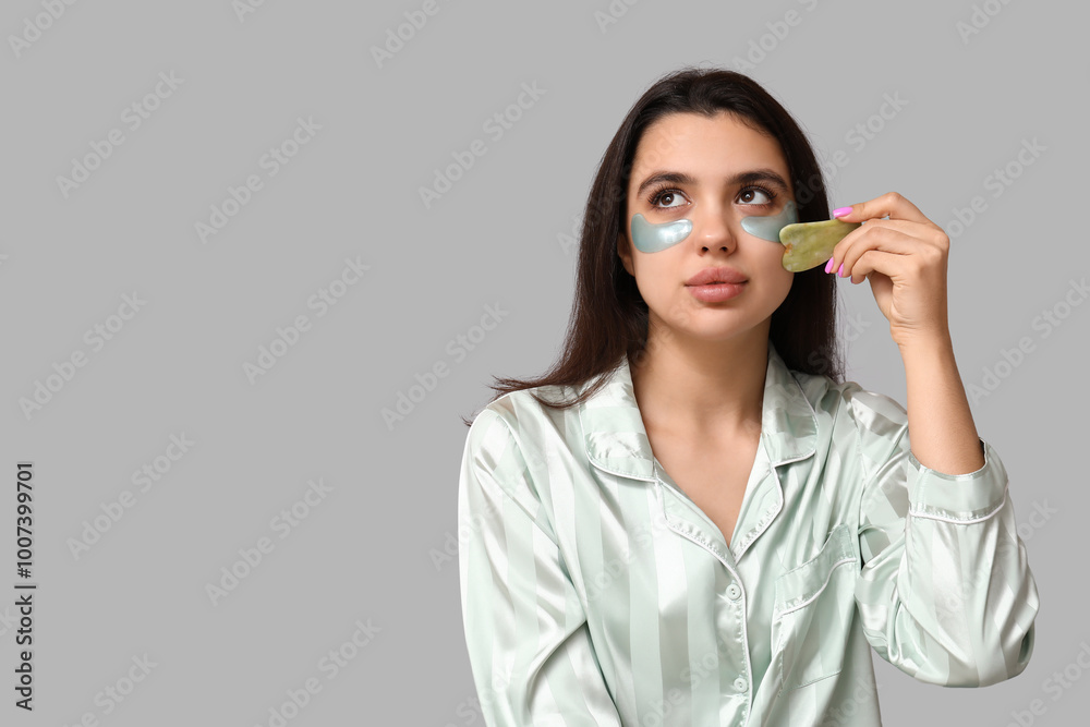 Beautiful young woman with under-eye patches using guasha massage tool on grey background Beautiful young woman with under-eye patches using guasha massage tool on grey background