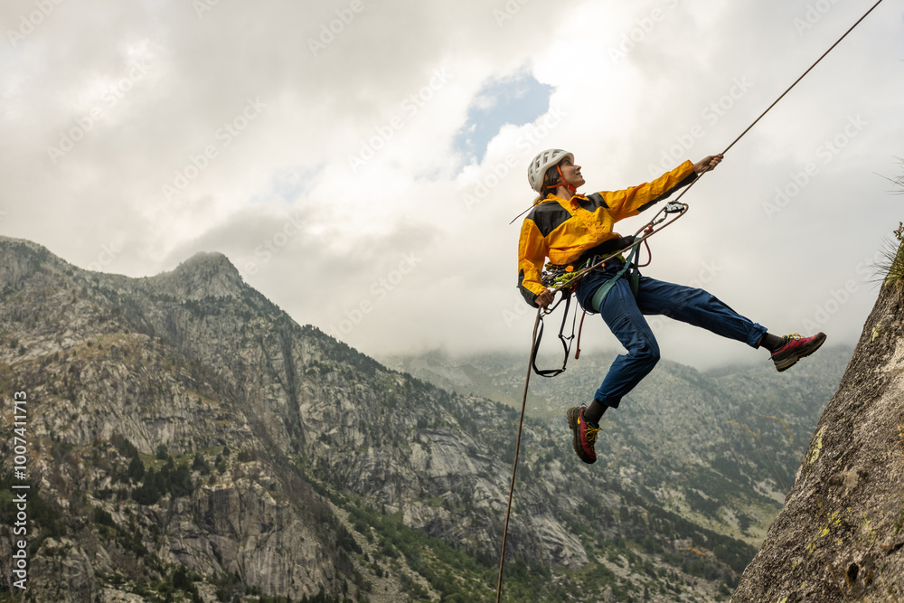 Obraz premium A man is jumping off a cliff with a rope attached to him. The man is wearing a yellow jacket and blue jeans. The sky is cloudy and the mountains are in the background. The man is wearing a helmet