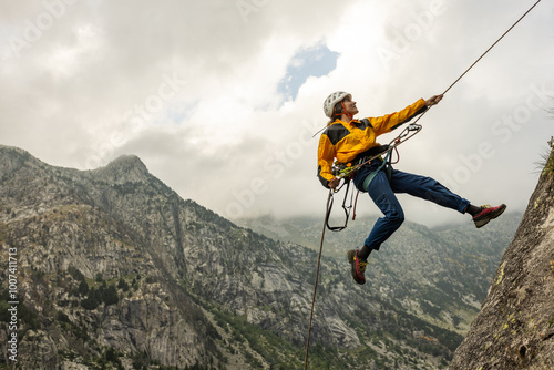 Fototapeta Naklejka Na Ścianę i Meble -  A man is jumping off a cliff with a rope attached to him. The man is wearing a yellow jacket and blue jeans. The sky is cloudy and the mountains are in the background. The man is wearing a helmet