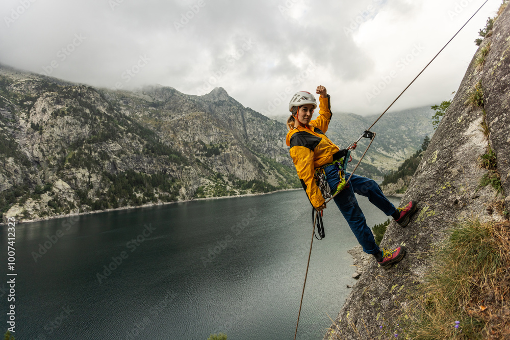 Obraz premium A woman is standing on a rock with a rope around her waist. She is wearing a yellow jacket and a helmet. The scene is set in a mountainous area with a lake in the background