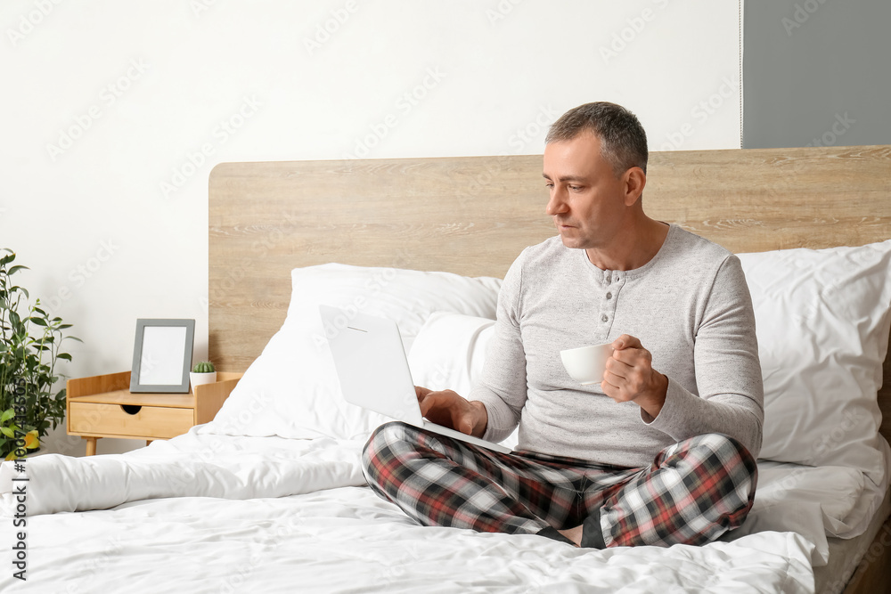Morning of handsome mature man in stylish pajamas with laptop drinking coffee on bed at home Morning of handsome mature man in stylish pajamas with laptop drinking coffee on bed at home
