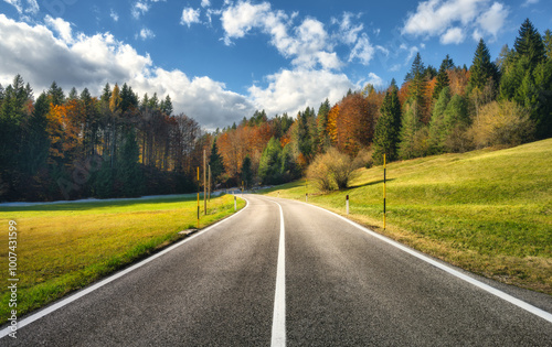 Road, green meadows and orange forest at sunset in golden autumn. Italy. Beautiful mountain road, trees, grass, blue sky with clouds. Landscape with empty highway, field and woods in fall. Travel