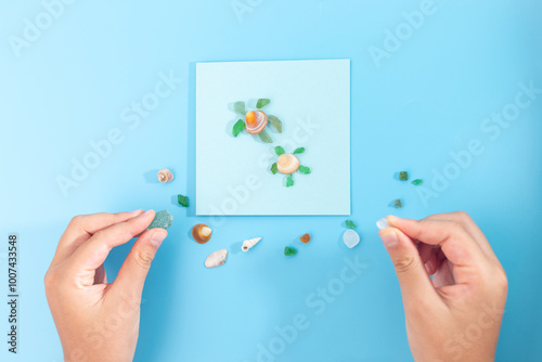 Artistic flat lay composition on blue background, hands adjusting shells, turtle made of shell craft, creating miniature beach scene, kids art project, nature materials, DIY, summer time vibe photo