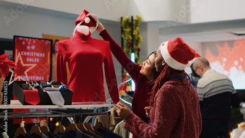 African american retail assistant wearing Santa hat helping asian client browsing through festive clothing in Christmas shopping store during winter holiday season. Employee assisting customer photo