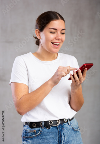 Portrait of cheerful young woman looking gladly at mobile phone against light unicoloured background photo