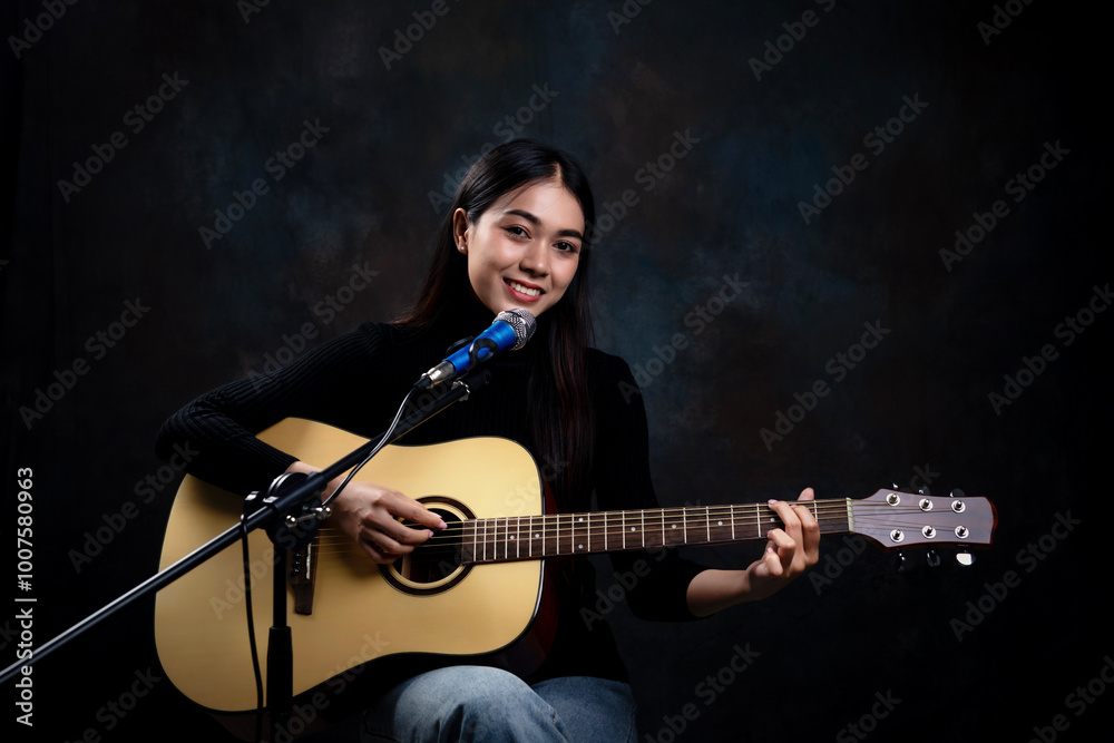 Obraz premium Beautiful young woman is playing the guitar and singing against a black background. Female playing guitar with passion and happiness