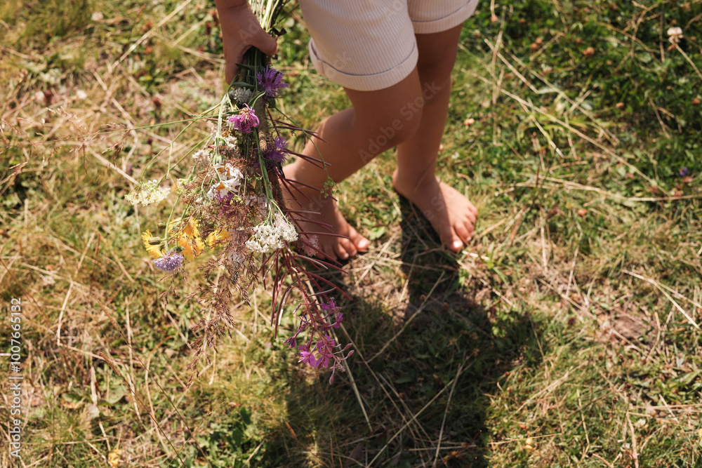 Fototapeta premium Little girl with bouquet of wildflowers walking barefoot outdoors on sunny day, closeup. Child enjoying beautiful nature