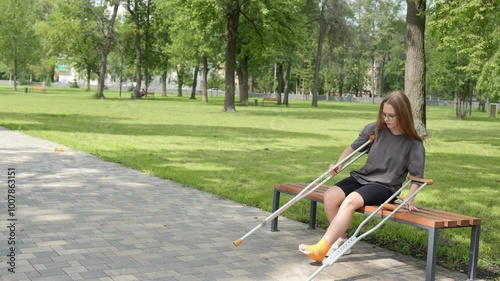 young woman with her leg in a cast carefully rises from a park bench, using crutches to support her weight as she navigates. photo