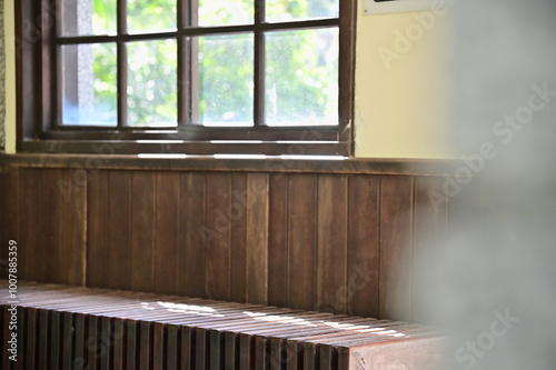 Soft sunlight passes through the window of a wooden Japanese-style train station, casting gentle shadows on the interior chairs. The light highlights the outdoor greenery, creating a peaceful ambiance