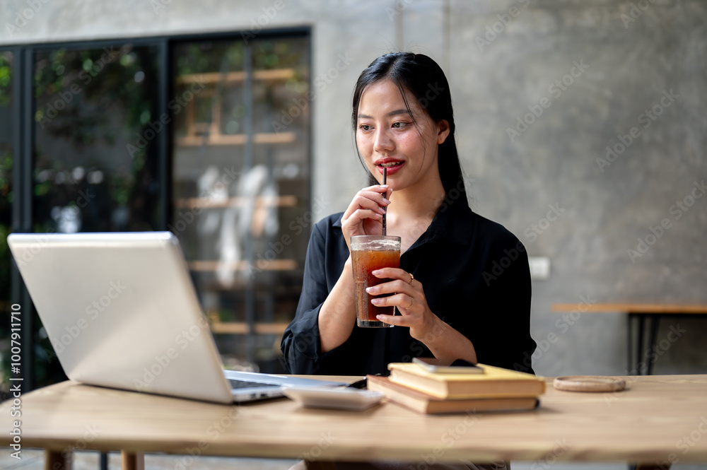 A beautiful Asian businesswoman enjoys iced coffee while working on her laptop, working remotely. A beautiful Asian businesswoman enjoys iced coffee while working on her laptop, working remotely.