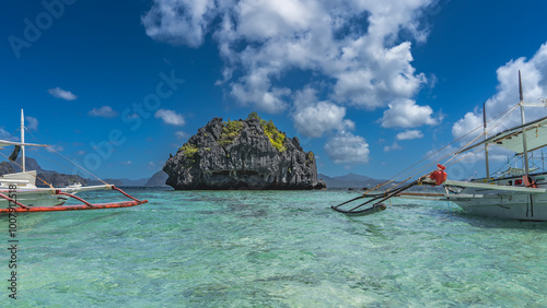 Traditional Filipino bangka boats are anchored in the ocean. Aquamarine water is crystal clear. A picturesque rocky island in the distance. Green vegetation on steep slopes. Blue sky, clouds.  photo